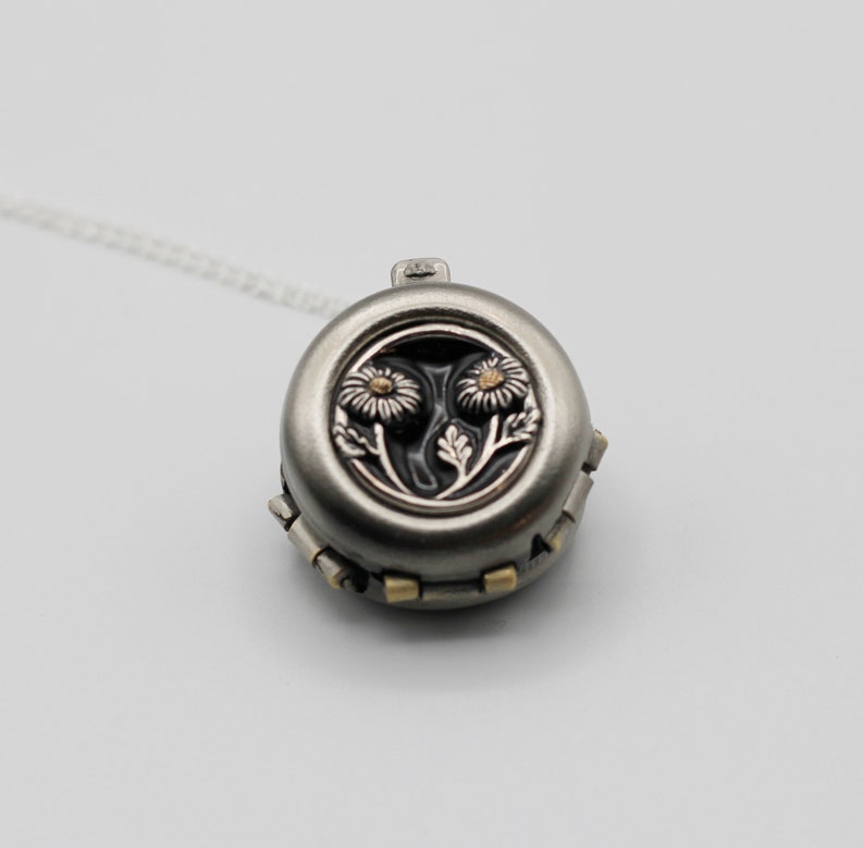 Peut inclure: Un collier pendentif rond argent&eacute; avec un motif floral. Le pendentif pr&eacute;sente deux fleurs d&eacute;taill&eacute;es sur un fond sombre. Le collier a une cha&icirc;ne d&eacute;licate et est pos&eacute; sur un fond blanc.