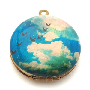 May include: A round, gold locket with a blue and green sky and white clouds painted on the front. Silhouettes of birds are flying across the sky.