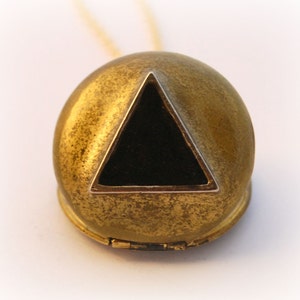 May include: A gold-toned pendant necklace with a round, domed locket that opens to reveal a black triangle inside. The locket has a worn, antique finish.