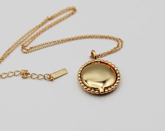 May include: A gold-toned necklace featuring a round, smooth pendant with a beaded edge. The chain is a delicate link style and includes a small rectangular tag near the clasp. The metal has a polished, reflective finish throughout the piece.
