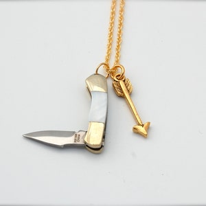 May include: A gold chain necklace with a small pocket knife and arrow charm. The knife has a white and gold handle and a silver blade. The arrow charm is gold and has a pointed tip.