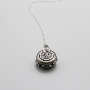 May include: A silver locket necklace with a compass design on the front. The locket is round and has a detailed design.