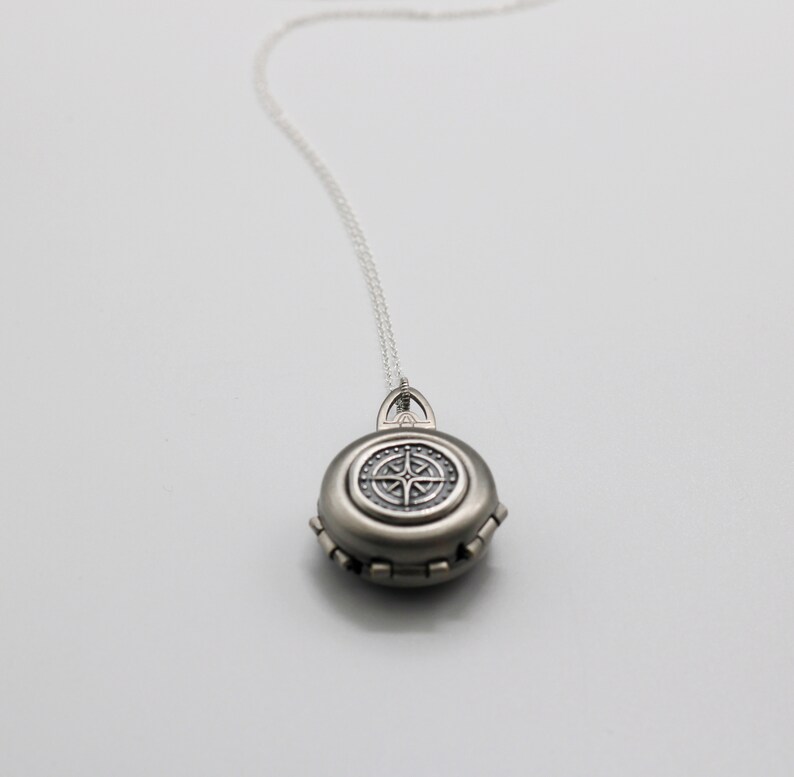 May include: A silver locket necklace with a compass design on the front. The locket is round and has a detailed design.