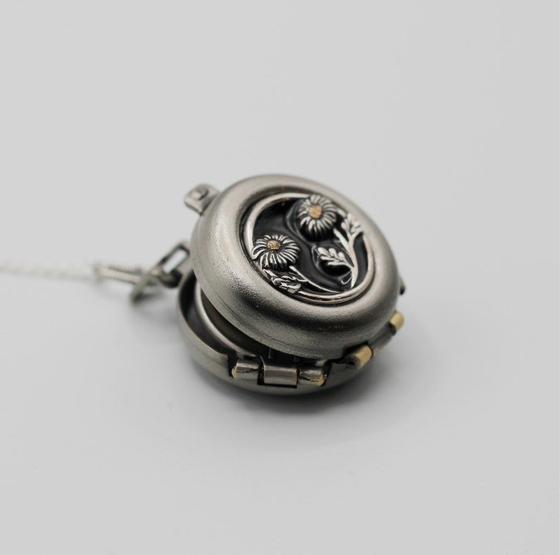 Peut inclure: Pendentif locket argent&eacute; ouvert avec un motif floral. Le locket pr&eacute;sente une gravure d&eacute;taill&eacute;e de deux marguerites avec des centres dor&eacute;s sur un fond noir. Le pendentif est attach&eacute; &agrave; une fine cha&icirc;ne argent&eacute;e.