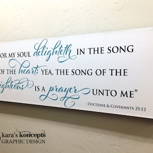 May include: White wooden sign with teal blue calligraphy text that reads "For my soul delighteth in the song of the heart; yea, the song of the righteous is a prayer unto me" - Doctrine & Covenants 25:12