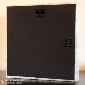 May include: The back of a framed canvas print with two hanging hardware pieces attached. The canvas is black and the frame is white with a distressed look.