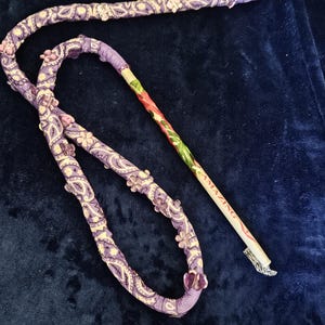 May include: A purple and white patterned fabric hose with a wooden stem and a white tip. The hose is decorated with purple and white flowers and the stem has the word "AMAZING" written on it.