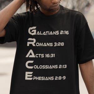 Könnte beinhalten: Ein schwarzes kurzärmeliges T-Shirt mit dem vertikal aufgedruckten Wort GRACE in großen, hellen Buchstaben. Jeder Buchstabe beginnt eine Textzeile mit Verweisen auf Bibelverse: Galatians 2:16, Romans 3:28, Acts 16:31, Colossians 2:13 und Ephesians 2:8-9.