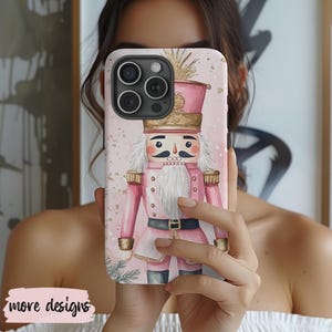 May include: A pink phone case featuring a nutcracker soldier design. The case has a light pink background with gold accents and a detailed illustration of the nutcracker in a pink uniform and a white beard. The text "more designs" is at the bottom.