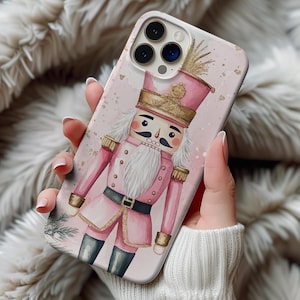 May include: A pink phone case featuring a watercolor illustration of a nutcracker soldier. The design includes gold accents, a white beard, and black boots. The case has a glossy finish and is held in a hand.