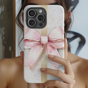 May include: White phone case featuring a watercolor design of a pink bow with gold accents. The case has a vertical gold stripe down the center and a speckled background. The phone case has a camera module with three lenses.