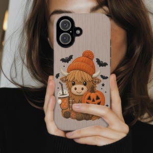 May include: A light beige phone case with a wood grain texture featuring a graphic of a brown highland cow wearing an orange knit beanie, holding an orange pumpkin and a beverage cup, surrounded by small black bats and stars.