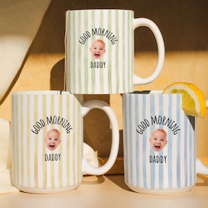 May include: Three ceramic mugs with vertical striped patterns in green, yellow, and blue. Each mug features the text "GOOD MORNING DADDY" with a baby's face. One mug has a lemon slice.
