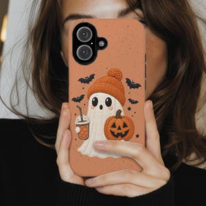 May include: A phone case with a speckled orange background featuring a white ghost wearing an orange knit beanie, holding a pumpkin and a beverage cup, surrounded by small black bats and stars, with a dual camera cutout at the top left corner.