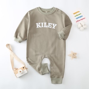 May include: A light green long-sleeved jumpsuit with a white embroidered name "KILEY" on the front. The jumpsuit has a snap closure at the crotch and snaps at the ankles.