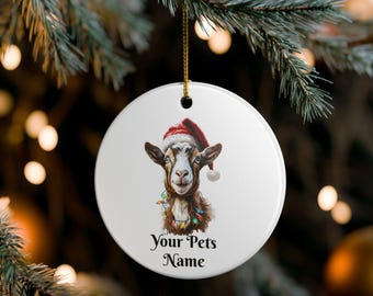 Könnte beinhalten: Ein weißer, runder Weihnachtsschmuck mit einer Ziege, die einen Weihnachtsmütze und eine Lichterkette trägt. Der Text "Your Pets Name" ist unter der Ziegenillustration aufgedruckt. Der Schmuck hängt an einem goldenen Faden vor einem verschwommenen Hintergrund aus Weihnachtslichtern.