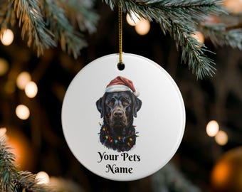 Könnte beinhalten: Ein weißer, runder Weihnachtsschmuck mit einem schwarzen Labrador Retriever, der einen Weihnachtsmannhut und eine Lichterkette trägt. Der Text "Your Pets Name" ist darunter gedruckt. Ein goldener Faden ist zum Aufhängen befestigt.