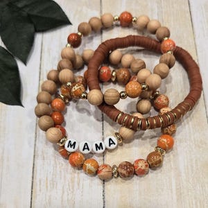 Op de afbeelding: Set van vier armbanden met oranje en bruine jaspis kralen, houten kralen en een bruin lederen armband met gouden accenten. De armbanden spellen het woord "MAMA" in witte letters.