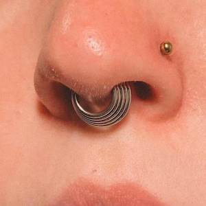 May include: A close-up shot of a face with multiple piercings. A silver, layered septum ring is visible, alongside a gold stud in the nostril and a lip piercing with a dangling silver ball. The word "lusi" is printed in black.