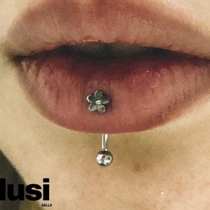 Può includere: Primo piano di un labbro inferiore con un piercing a barra a forma di fiore argentato. La barra ha una piccola sfera argentata sul fondo.