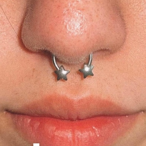 May include: Close-up of a person's face with a septum piercing featuring two silver star-shaped charms. The piercing is centered on the septum, with the stars facing outward.