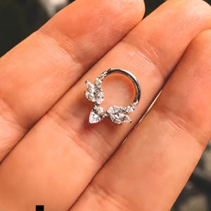 May include: A silver nose ring with a floral design. The ring features clear gemstones arranged in a leaf-like pattern. The ring is held in a person's hand.