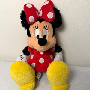 May include: A plush Minnie Mouse doll, with a red dress and white polka dots, a matching bow, and yellow shoes. The doll has black fur, white gloves, and a smiling face. A classic Disney character.