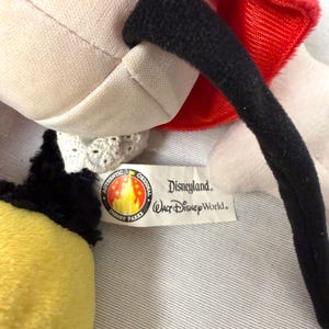 May include: A close-up of a plush toy with a red bow, black ear, and yellow and black section. The toy has a white body and a tag that reads "Disneyland Walt Disney World" and "Authentic Original Disney Parks."