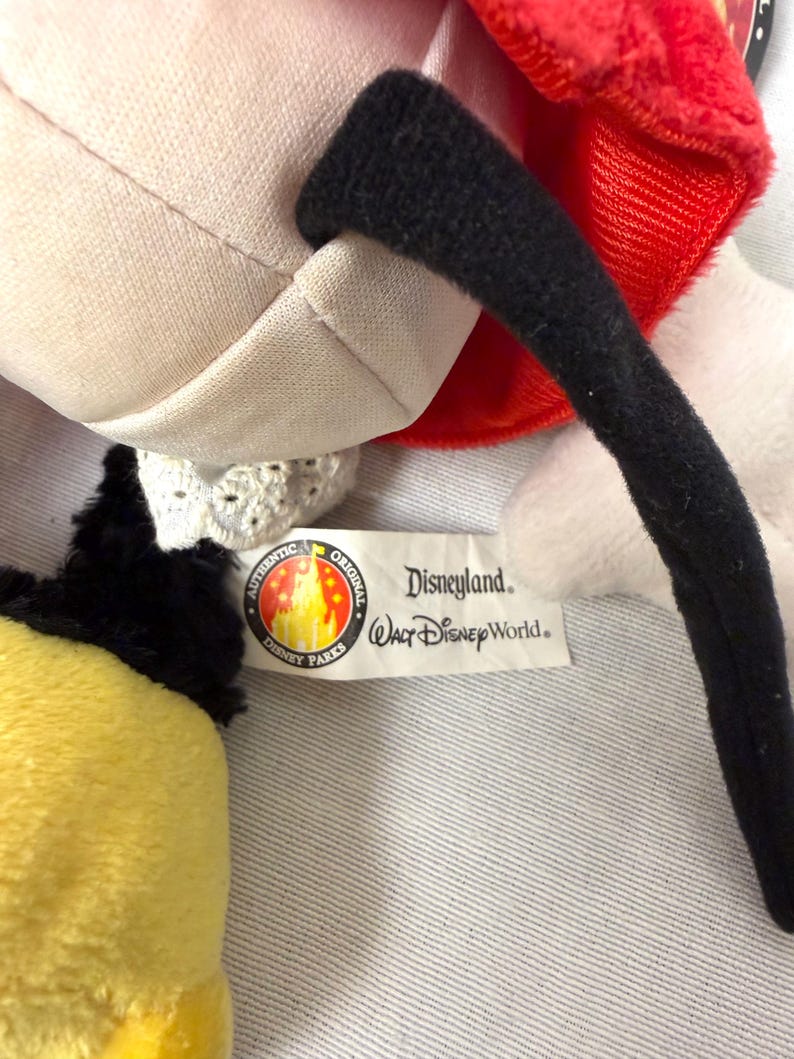 May include: A close-up of a plush toy with a red bow, black ear, and yellow and black section. The toy has a white body and a tag that reads "Disneyland Walt Disney World" and "Authentic Original Disney Parks."