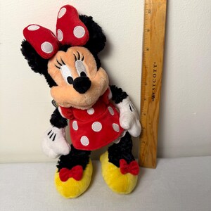 May include: A Minnie Mouse plush toy. The plush has a red dress with white polka dots, a red bow with white polka dots, and yellow shoes. The toy is approximately 25 cm tall.