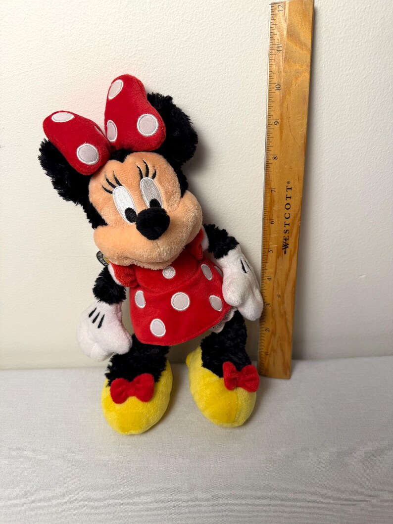 May include: A Minnie Mouse plush toy. The plush has a red dress with white polka dots, a red bow with white polka dots, and yellow shoes. The toy is approximately 25 cm tall.
