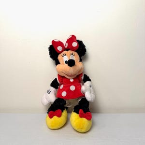 May include: A Minnie Mouse soft toy. She is dressed in a red dress with white polka dots, a matching bow, and yellow shoes with red bows. Her black fur is visible on her head, arms, and legs. Her white gloves and facial features are also visible.