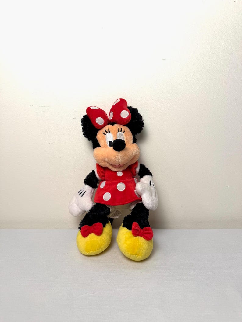 May include: A Minnie Mouse soft toy. She is dressed in a red dress with white polka dots, a matching bow, and yellow shoes with red bows. Her black fur is visible on her head, arms, and legs. Her white gloves and facial features are also visible.