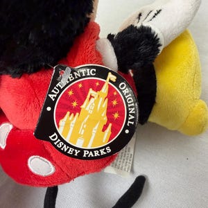 May include: A Mickey Mouse soft toy with a red body, black ears, and a white glove. The toy has a Disney Parks tag attached, featuring a castle graphic and the words "Authentic Original". The toy's tail is black.