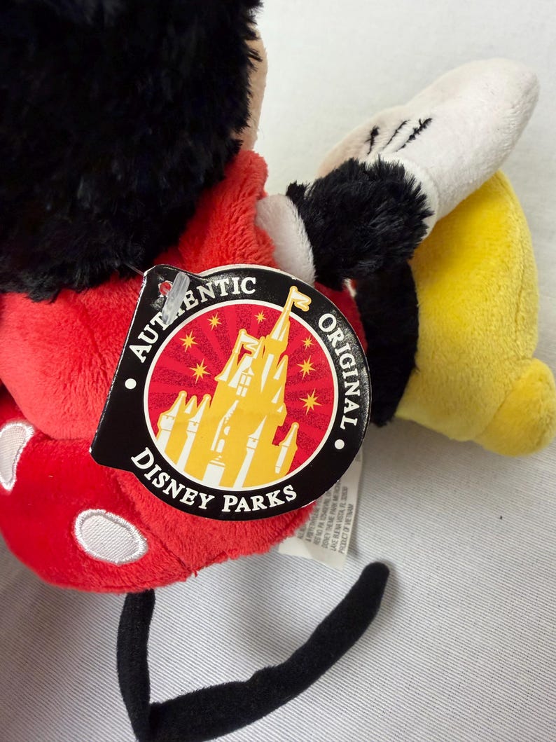 May include: A Mickey Mouse soft toy with a red body, black ears, and a white glove. The toy has a Disney Parks tag attached, featuring a castle graphic and the words "Authentic Original". The toy's tail is black.