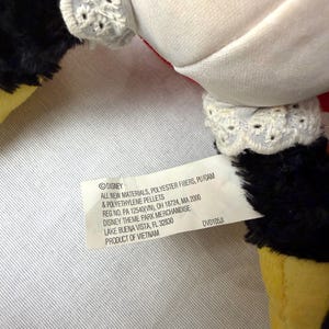 May include: Close-up of a plush toy with white fabric, black fur, and yellow feet. The toy has white lace trim and a tag with text including "DISNEY" and "PRODUCT OF VIETNAM".