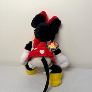 May include: A Minnie Mouse soft toy, seen from behind, wearing a red and white polka dot dress, a red bow, and yellow shoes. The black and white toy has a tail and white gloves. A Disney Parks tag is attached.