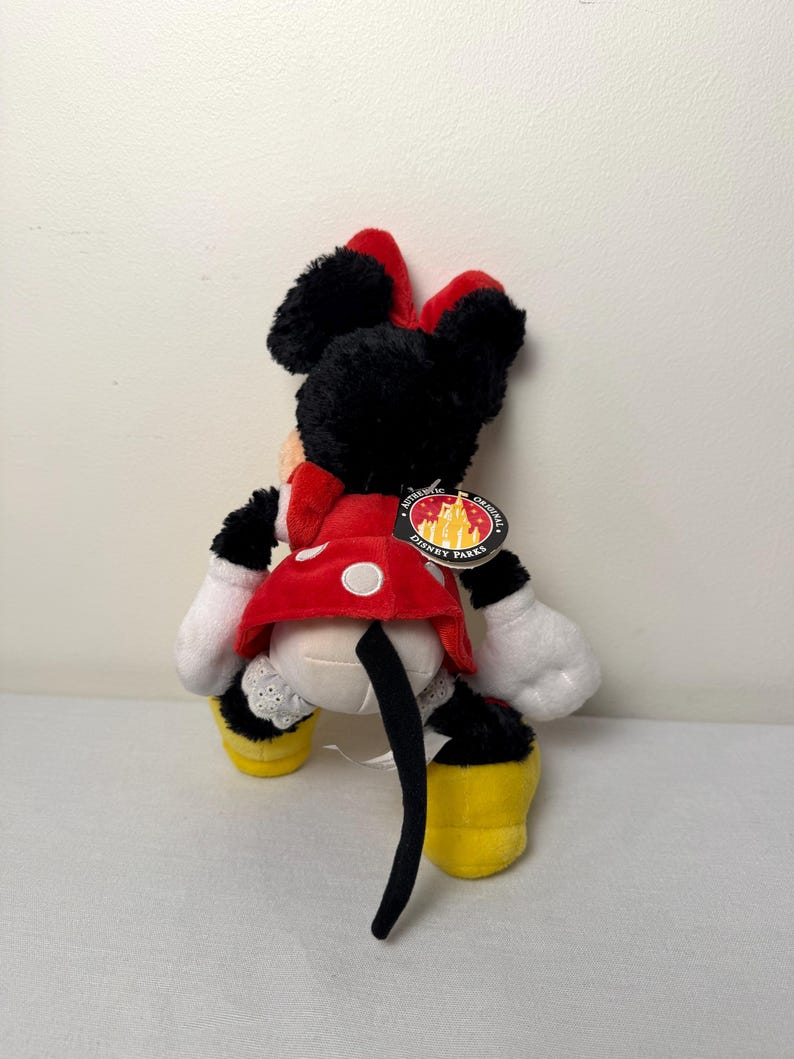 May include: A Minnie Mouse soft toy, seen from behind, wearing a red and white polka dot dress, a red bow, and yellow shoes. The black and white toy has a tail and white gloves. A Disney Parks tag is attached.