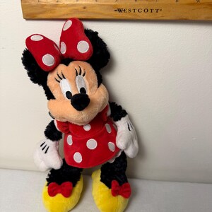 May include: A Minnie Mouse soft toy. The toy features a red dress with white polka dots, a red bow with white polka dots, and yellow shoes with red bows. The toy is black, red, white, and yellow.