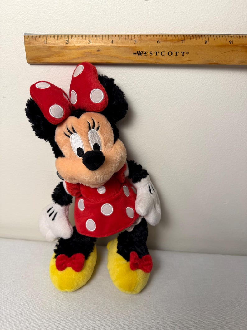 May include: A Minnie Mouse soft toy. The toy features a red dress with white polka dots, a red bow with white polka dots, and yellow shoes with red bows. The toy is black, red, white, and yellow.