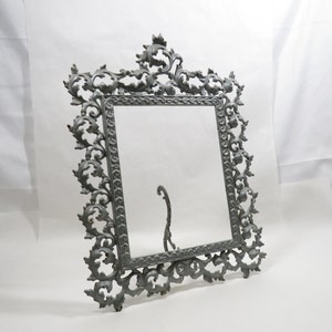 May include: A silver-colored metal picture frame with an ornate, scroll-like design. The frame has a stand on the back for displaying on a table.