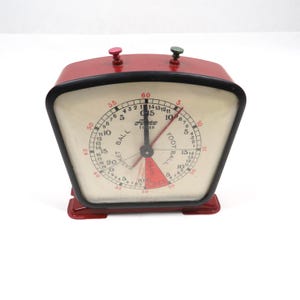 May include: A vintage red Auto Timer with a black trim and a cream-colored face. The timer features two knobs, a black hand, and a red and black segmented dial for basketball and football. The face displays numbers for minutes and seconds.