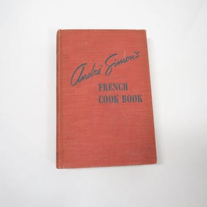 May include: A vintage cookbook with a red cover. The title reads "Andre Simon's FRENCH COOK BOOK" in black lettering. The book has a worn appearance, suggesting it is an antique or a well-loved item.