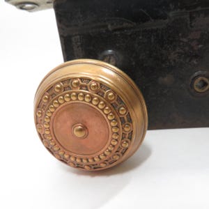 May include: A brass door knob with a decorative pattern around the edge and a copper center. The knob is round and has a smooth surface.