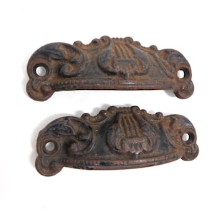 May include: Two antique cast iron drawer pulls with an ornate, scrollwork design. The pulls are dark brown with a distressed finish, featuring a central lyre motif and mounting holes on each end. These decorative hardware pieces are ideal for vintage furniture restoration.