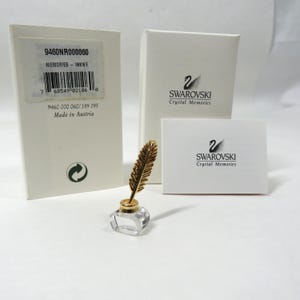 May include: A clear crystal inkwell with a gold-coloured feather pen. The inkwell is square-shaped. The image includes the original packaging with the text "SWAROVSKI Crystal Memories". The product is labeled "MEMORIES - INKWE".