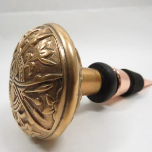 May include: A gold-toned bottle stopper with an ornate design. The stopper has a round, decorative top and a long, thin neck.