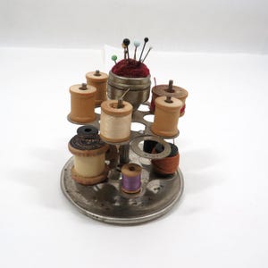 May include: A vintage metal thread stand with a pincushion on top. The stand has multiple spools of thread in various colors, including brown, white, purple, and orange. The pincushion is red with a metal base and has several colorful pins.