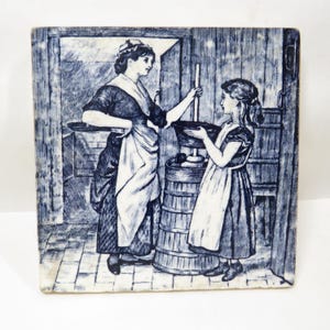 May include: Blue and white ceramic tile with a detailed illustration of two women in a kitchen. One woman holds a long wooden tool, the other a bowl. The scene evokes a historical period, with a rustic aesthetic.