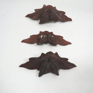 May include: Three dark brown, carved wooden decorative leaf and pod ornaments. Each ornament features a central pod shape with detailed leaf-like extensions. The ornaments are arranged on a white background.